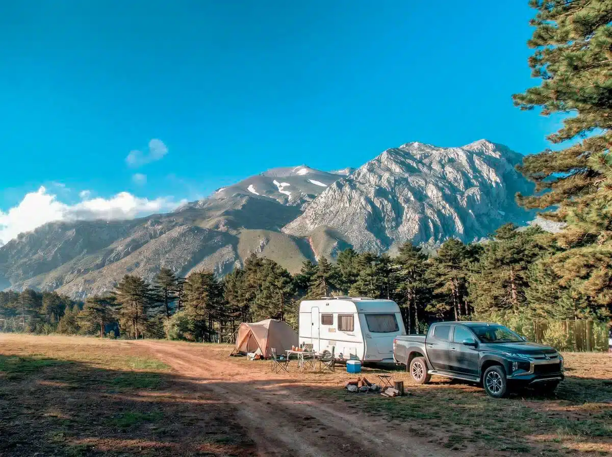 Toprak yol kenarında heybetli dağ manzarasına karşı park etmiş pickup arazi aracı, çekme karavan ve kamp çadırı.
