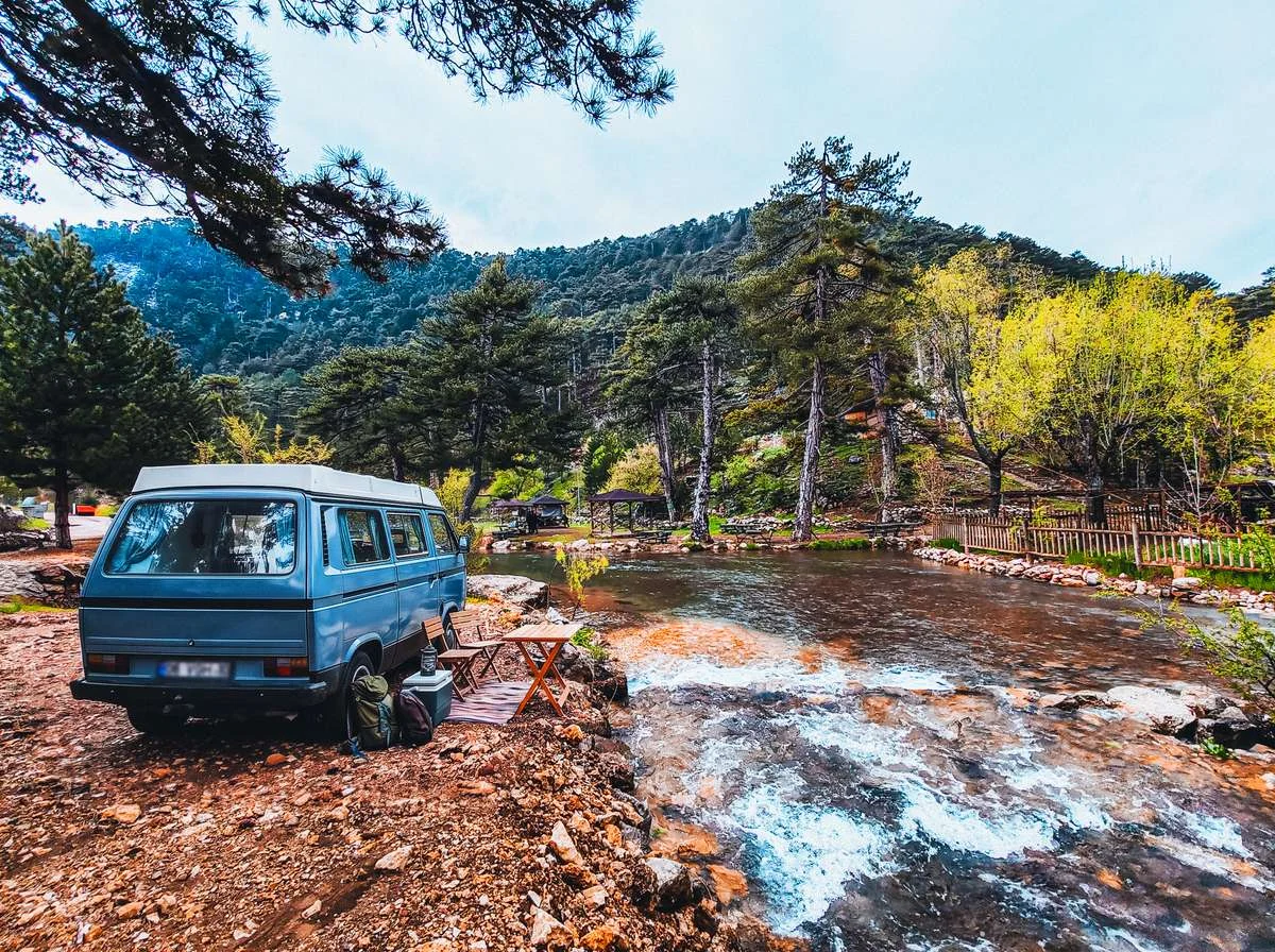 Başpınar Tabiat Parkı'nda dere kenarına park etmiş mavi bir karavan ve dışarıya kurulmuş portatif masalı dinlenme alanı.
