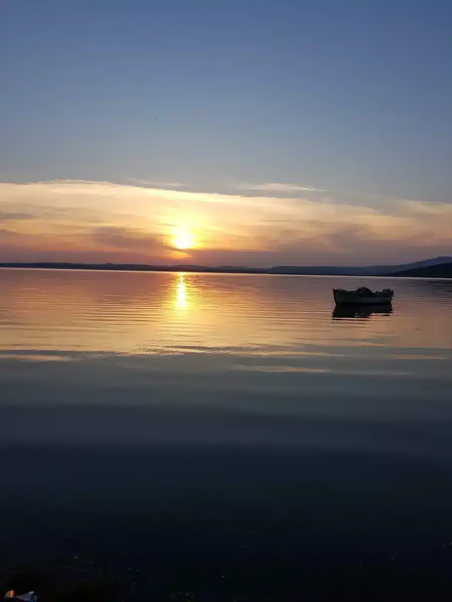 Akçalar kıyısından Uluabat Gölü üzerinde gün batımı kızıllığı, durgun su yansıması ve göldeki küçük sandal.